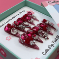 Bougie Bling 11 press on nails by CutiePop Nail Shop featuring long stiletto shaped nails with a bold wine red glitter base layered with luxurious oversized 3D gem clusters in deep jewel tones, accented with sparkling rhinestones and metallic detailing. Hand sculpted for a dramatic, upscale statement mani.