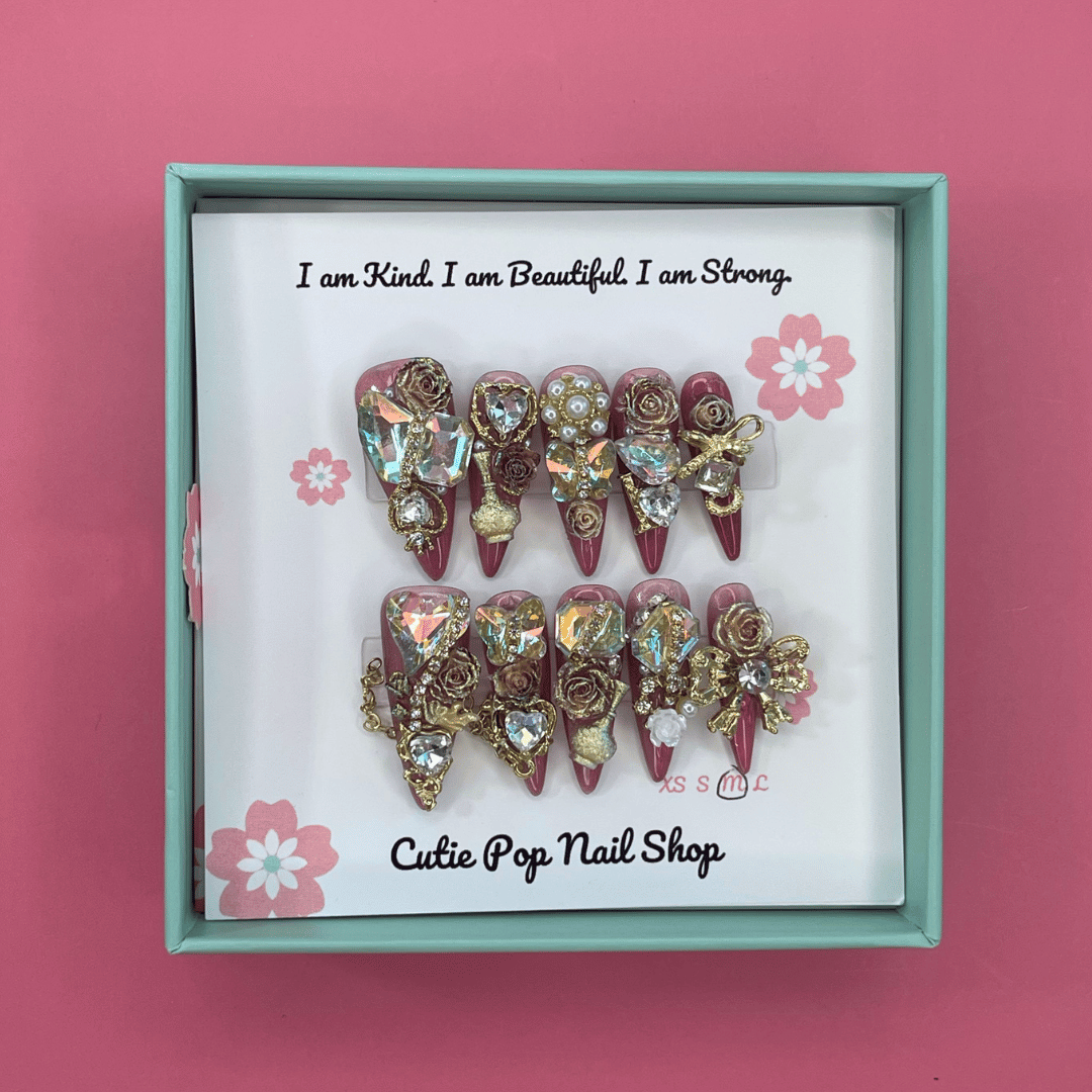 Bougie Bling 12 press on nails by CutiePop Nail Shop featuring long stiletto shaped nails with a glamorous deep dark burgundy or smoky wine toned glitter base. Lavish oversized 3D gem clusters in jewel tones such as ruby and crystal are complemented by sparkling rhinestones and metallic accents—arranged in an opulent, high impact luxury statement manicure.

