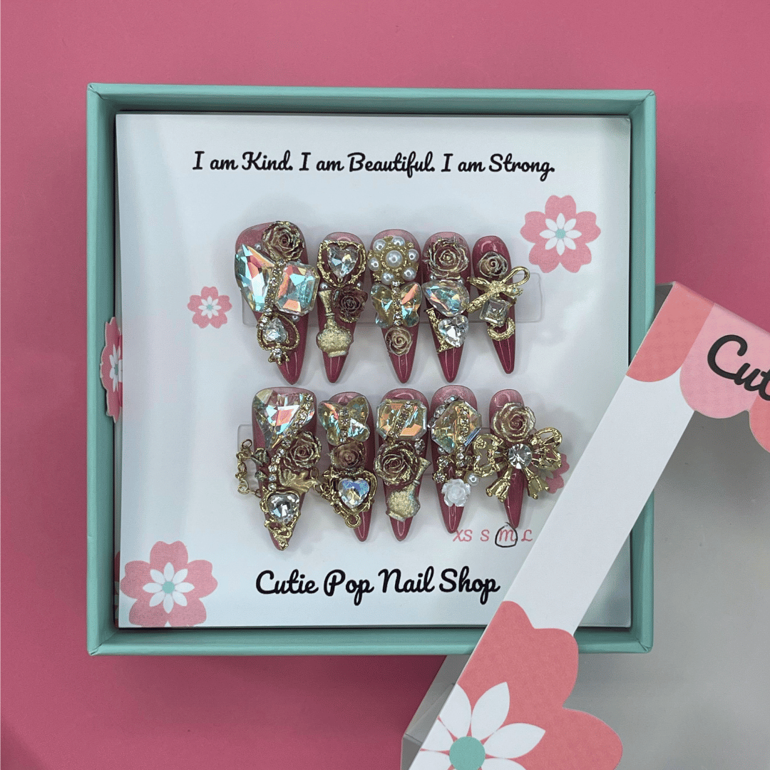 Bougie Bling 12 press on nails by CutiePop Nail Shop featuring long stiletto shaped nails with a glamorous deep dark burgundy or smoky wine toned glitter base. Lavish oversized 3D gem clusters in jewel tones such as ruby and crystal are complemented by sparkling rhinestones and metallic accents—arranged in an opulent, high impact luxury statement manicure.

