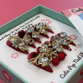 Bougie Bling 13 press on nails by CutiePop Nail Shop featuring long stiletto shaped nails with a rich deep burgundy red or wine toned glitter base. Lavish oversized 3D gem clusters in jewel tones including ruby and crystal are accentuated with sparkling rhinestones and metallic highlights for a dramatic, high end glitzy manicure.