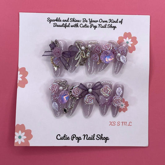 Bougie Bling 16 press-on nails by CutiePop Nail Shop feature medium-length almond-shaped nails in a shimmering lavender-purple glitter base, adorned with ornate 3D gem clusters and delicate rhinestone accents. Each nail is meticulously hand-sculpted to create a luxurious, playful-glam statement manicure. Includes 10 reusable nails, jelly adhesive tabs, a mini nail file, and a cuticle stick.