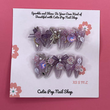 Bougie Bling 16 press-on nails by CutiePop Nail Shop feature medium-length almond-shaped nails in a shimmering lavender-purple glitter base, adorned with ornate 3D gem clusters and delicate rhinestone accents. Each nail is meticulously hand-sculpted to create a luxurious, playful-glam statement manicure. Includes 10 reusable nails, jelly adhesive tabs, a mini nail file, and a cuticle stick.

