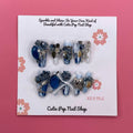 Bougie Bling 17 press‑on nails by CutiePop Nail Shop featuring medium-length coffin-shaped nails with a deep navy or black glitter base, paired with lavish oversized 3D gem clusters in complementary jewel tones. Sparkling rhinestone accents and metallic touches complete this dramatic, high‑glam statement manicure.

