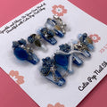 Bougie Bling 17 press‑on nails by CutiePop Nail Shop featuring medium-length coffin-shaped nails with a deep navy or black glitter base, paired with lavish oversized 3D gem clusters in complementary jewel tones. Sparkling rhinestone accents and metallic touches complete this dramatic, high‑glam statement manicure.

