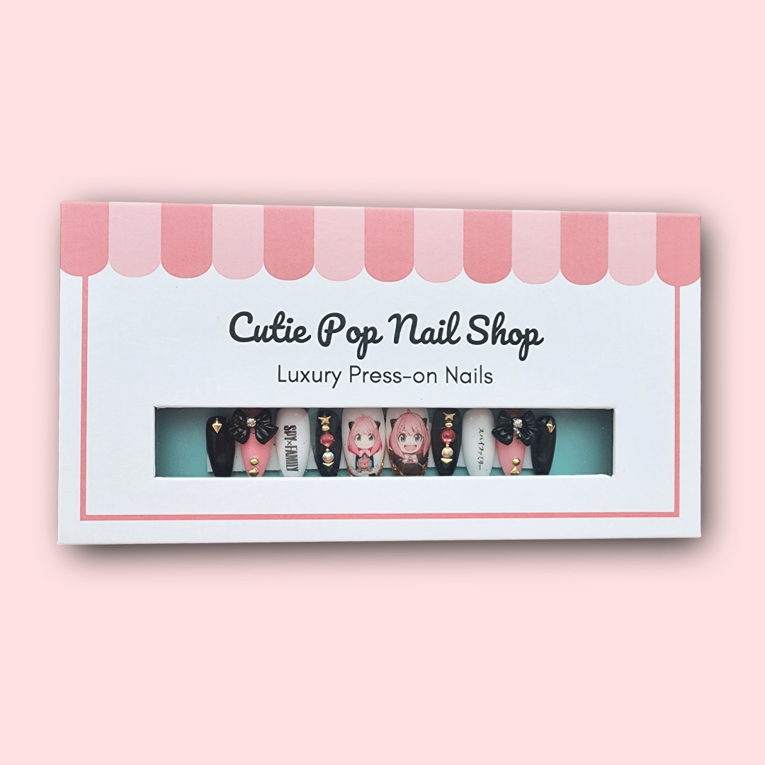 Anya press‑on nails by CutiePop Nail Shop featuring medium-length almond-shaped nails in a mix of pastel pink, lavender, and black tones. Each nail showcases playful chibi-style decals of Anya from Spy × Family, accented with tiny hearts, stars, and glossy gel shine. The set includes 10 handmade reusable nails, jelly adhesive tabs, a mini nail file, and a cuticle stick—crafted for fans of kawaii anime style and covert charm.