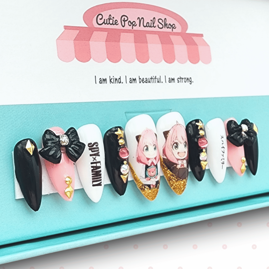 Anya press‑on nails by CutiePop Nail Shop featuring medium-length almond-shaped nails in a mix of pastel pink, lavender, and black tones. Each nail showcases playful chibi-style decals of Anya from Spy × Family, accented with tiny hearts, stars, and glossy gel shine. The set includes 10 handmade reusable nails, jelly adhesive tabs, a mini nail file, and a cuticle stick—crafted for fans of kawaii anime style and covert charm.
