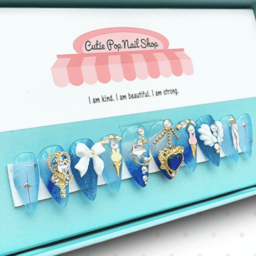 Mercury press on nails by CutiePop Nail Shop featuring medium stiletto shaped nails in pastel aqua blue and soft white tones. Several accent nails highlight hand painted chibi-style Princess Mercury illustrations, crescent moon decals, holo-flake glitter, and water droplet motifs. Finished with a glossy gel coat and metallic shimmer that capture Sailor Mercury’s graceful, aquatic elegance. The set includes 10 reusable handmade nails, jelly adhesive tabs, mini nail file, and cuticle stick.
