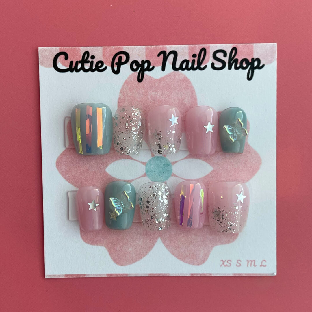Dreamy Butterfly press‑on nails by CutiePop Nail Shop featuring coffin-shaped extra short nails in soft pastel pinks and blues, accented with hand-painted butterfly wing motifs, metallic foil shimmer, and delicate glitter sparkle. The set includes reusable press-on tabs, a mini nail file, and a cuticle stick—offering a fantasy-inspired, playful yet elegant manicure.