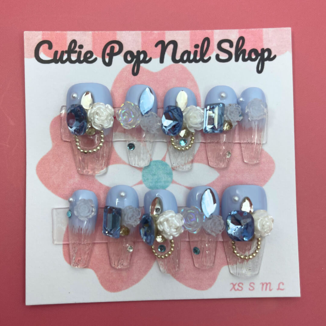 Elegant Rose (Blue) press on nails by CutiePop Nail Shop featuring medium coffin shaped nails in a rich sapphire blue or teal base, decorated with finely hand painted rose motifs in soft blush or pale pink tones. Accented with subtle metallic shimmer and tiny pearl or crystal details at the rose centers, finished with a glossy gel top coat. The set includes 10 reusable handmade nails, jelly adhesive tabs, a mini nail file, and a cuticle stick—designed for a refined, floral luxe manicure.

