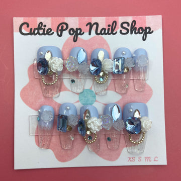 Elegant Rose (Blue) press on nails by CutiePop Nail Shop featuring medium coffin shaped nails in a rich sapphire blue or teal base, decorated with finely hand painted rose motifs in soft blush or pale pink tones. Accented with subtle metallic shimmer and tiny pearl or crystal details at the rose centers, finished with a glossy gel top coat. The set includes 10 reusable handmade nails, jelly adhesive tabs, a mini nail file, and a cuticle stick—designed for a refined, floral luxe manicure.

