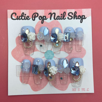 Elegant Rose (Blue) press‑on nails by CutiePop Nail Shop featuring long coffin-shaped nails in a rich sapphire-blue or teal base, decorated with finely hand-painted rose motifs in soft blush or pale pink tones. Accented with subtle metallic shimmer and tiny pearl or crystal details at rose centers, finished with a glossy gel top coat. The set includes 10 reusable handmade nails, jelly adhesive tabs, a mini nail file, and a cuticle stick—designed for a refined, floral-luxe manicure.