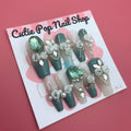 Elegant Rose (Green) press‑on nails by CutiePop Nail Shop featuring long coffin-shaped nails in a rich emerald-green base accented with intricately hand-painted delicate rose motifs in soft beige or cream hues. Embellished with subtle metallic shimmer and tiny pearl or rhinestone details at the center of each rose, finished with glossy gel polish for a refined, elegant floral manicure. Includes 10 handmade reusable nails, jelly adhesive tabs, a mini nail file, and cuticle stick for a full DIY kit.