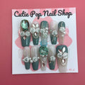 Elegant Rose (Green) press‑on nails by CutiePop Nail Shop featuring long coffin-shaped nails in a rich emerald-green base accented with intricately hand-painted delicate rose motifs in soft beige or cream hues. Embellished with subtle metallic shimmer and tiny pearl or rhinestone details at the center of each rose, finished with glossy gel polish for a refined, elegant floral manicure. Includes 10 handmade reusable nails, jelly adhesive tabs, a mini nail file, and cuticle stick for a full DIY kit.