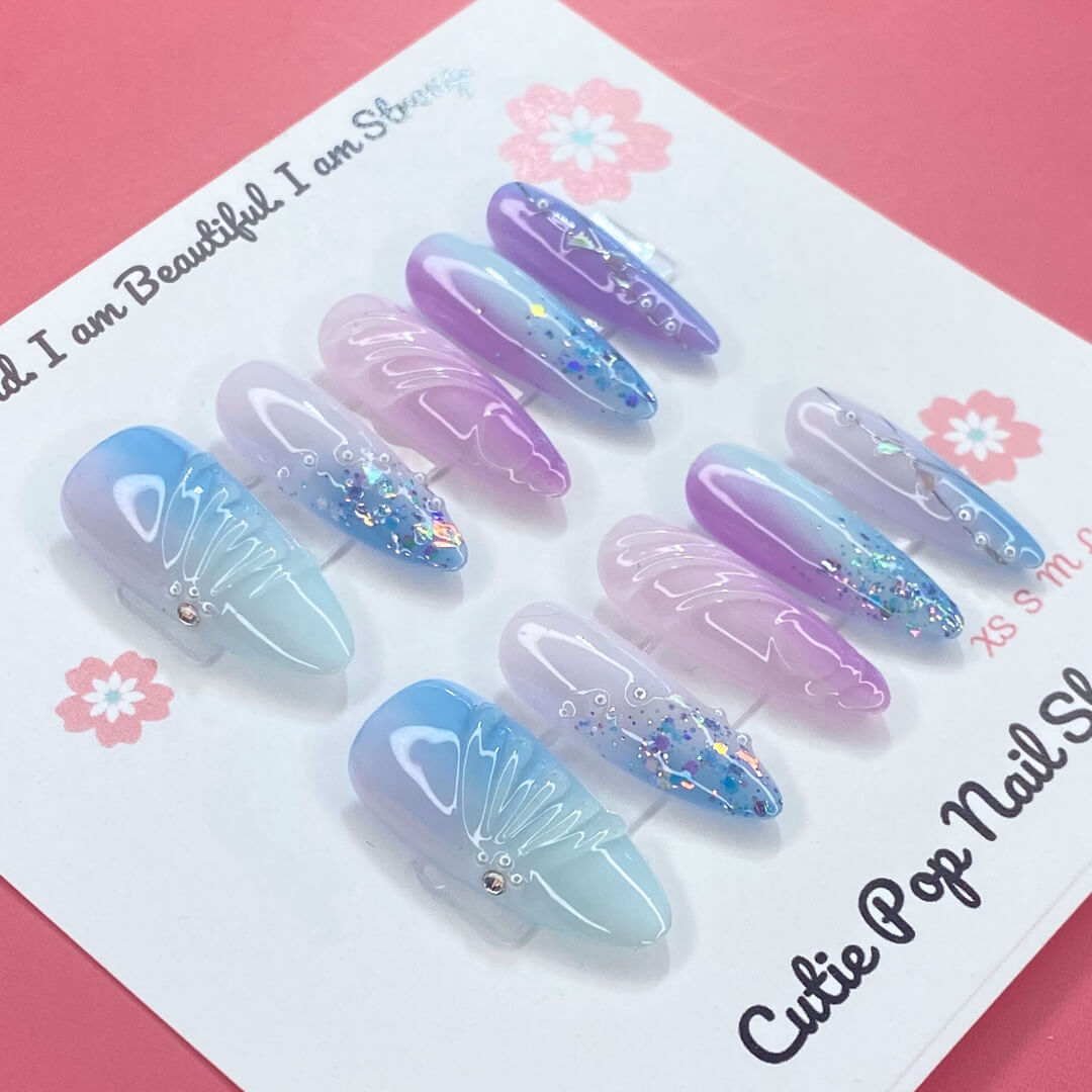Enchanted Ombre press on nails by CutiePop Nail Shop featuring long stiletto shaped nails with a dreamy pastel ombré blend of icy blue, lavender, and soft pink tones. Accented with chunky glitter, sparkling gems, and intricate embossed details—finished in a glossy coat for a magical, whimsical aesthetic.

