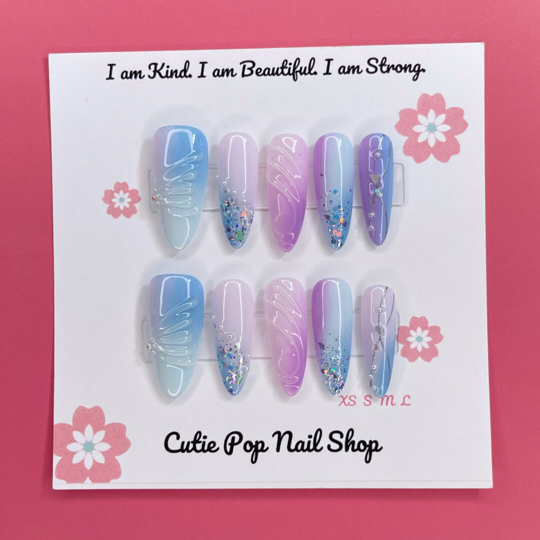 Enchanted Ombre press on nails by CutiePop Nail Shop featuring long stiletto shaped nails with a dreamy pastel ombré blend of icy blue, lavender, and soft pink tones. Accented with chunky glitter, sparkling gems, and intricate embossed details—finished in a glossy coat for a magical, whimsical aesthetic.

