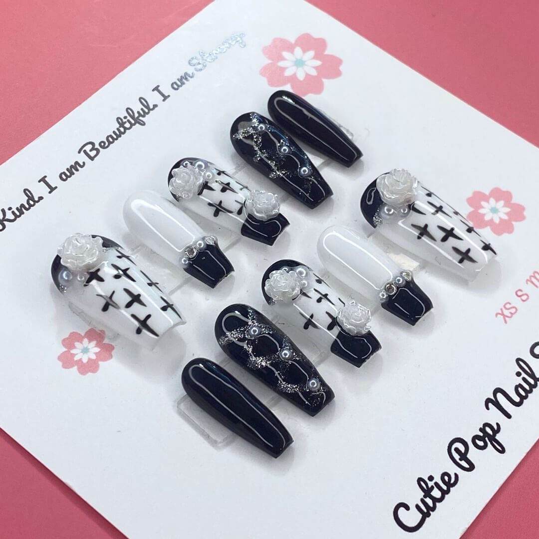 Floral Cross Monochrome press‑on nails by CutiePop Nail Shop featuring a chic coffin-shaped medium-length set in a classic black-and-white palette. Hand-painted white flowers and bold black crosses decorate alternating nails, accentuated by subtle metallic shimmer and glossy gel finish—creating a sophisticated gothic-inspired floral aesthetic.