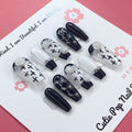 Floral Cross Monochrome press‑on nails by CutiePop Nail Shop featuring a chic coffin-shaped medium-length set in a classic black-and-white palette. Hand-painted white flowers and bold black crosses decorate alternating nails, accentuated by subtle metallic shimmer and glossy gel finish—creating a sophisticated gothic-inspired floral aesthetic.