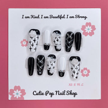 Floral Cross Monochrome press‑on nails by CutiePop Nail Shop featuring a chic coffin-shaped medium-length set in a classic black-and-white palette. Hand-painted white flowers and bold black crosses decorate alternating nails, accentuated by subtle metallic shimmer and glossy gel finish—creating a sophisticated gothic-inspired floral aesthetic.