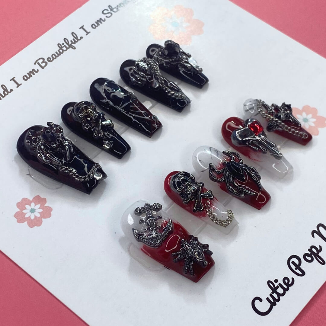 Gothic Delight press on nails by CutiePop Nail Shop featuring long coffin shaped nails painted in glossy jet black polish, accented with dark red duochrome shimmer and intricate gothic motifs like crosses or rose patterns, finished with subtle metallic or glitter highlights for a dramatic, dark romantic aesthetic.
