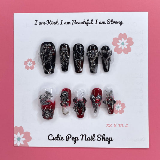 Gothic Delight press on nails by CutiePop Nail Shop featuring long coffin shaped nails painted in glossy jet black polish, accented with dark red duochrome shimmer and intricate gothic motifs like crosses or rose patterns, finished with subtle metallic or glitter highlights for a dramatic, dark romantic aesthetic.