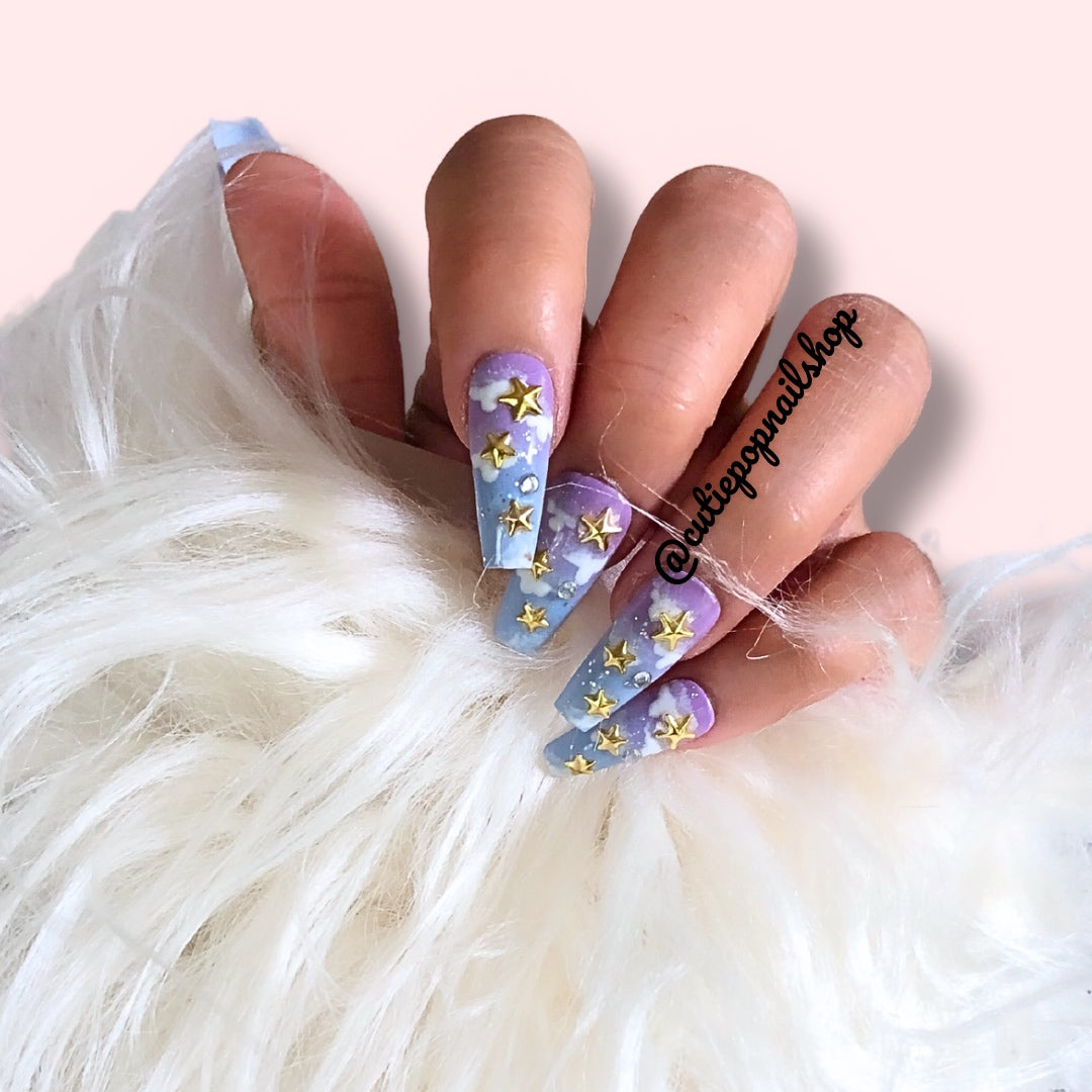 Twilight press‑on nails by CutiePop Nail Shop featuring medium-length coffin-shaped nails in a gradient blend of deep midnight blue fading into dusty purple or black. Accented with silver metallic star motifs, tiny moon charms, and scattered holographic shimmer, finished with a high-gloss gel polish. The set includes ten reusable nails, jelly adhesive tabs, a mini nail file, and a cuticle stick—designed for a dreamy, celestial manicure evoking twilit magic.

