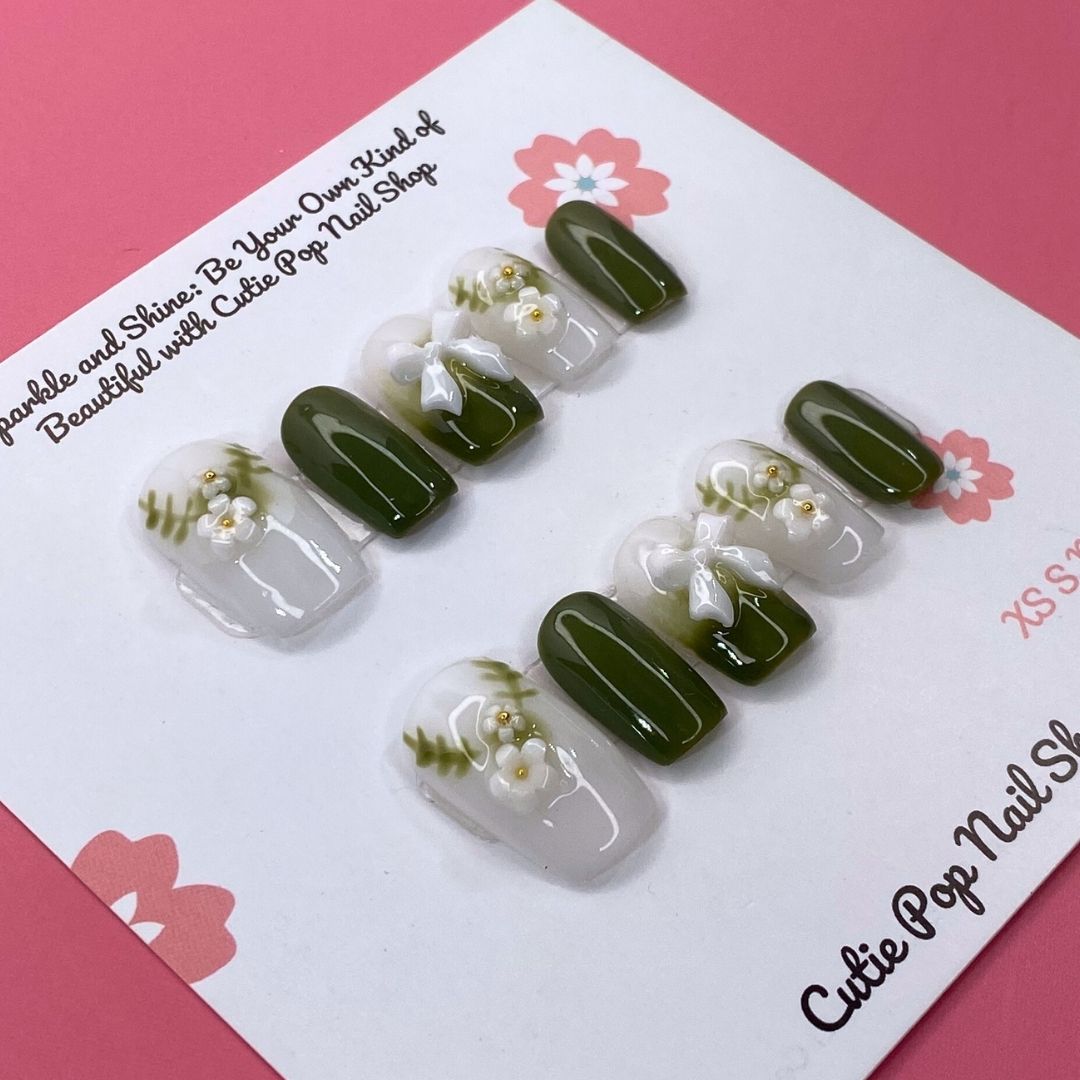 Lovely Olive press on nails by CutiePop Nail Shop featuring short round shaped nails in a soft white and olive green color palette. Adorned with delicate ornaments and charming ribbon accents, this design offers a sweet, festive-inspired look with a clean and elegant finish.

