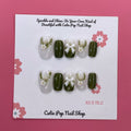 Lovely Olive press on nails by CutiePop Nail Shop featuring short round shaped nails in a soft white and olive green color palette. Adorned with delicate ornaments and charming ribbon accents, this design offers a sweet, festive-inspired look with a clean and elegant finish.

