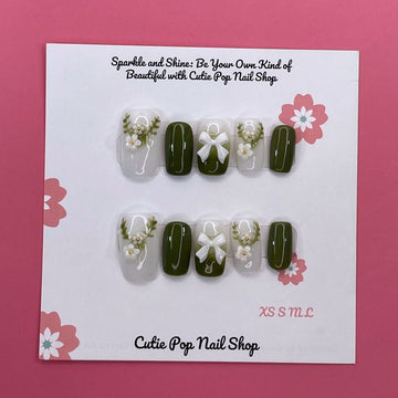 Lovely Olive press on nails by CutiePop Nail Shop featuring short round shaped nails in a soft white and olive green color palette. Adorned with delicate ornaments and charming ribbon accents, this design offers a sweet, festive-inspired look with a clean and elegant finish.

