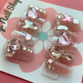 Magical Rhinestones press‑on nails by CutiePop Nail Shop featuring short to medium-length coffin-shaped nails in a soft blush-nude base, each adorned with oversized, iridescent rhinestone clusters forming floral and starburst patterns. The design sparkles with multi-faceted gem accents and metallic highlights for a luxe, high-bling aesthetic. The kit includes 10 reusable handmade nails, jelly adhesive tabs, a mini nail file, and a cuticle stick—perfect for glamorous DIY application.
