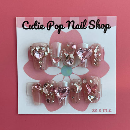 Magical Rhinestones press‑on nails by CutiePop Nail Shop featuring short to medium-length coffin-shaped nails in a soft blush-nude base, each adorned with oversized, iridescent rhinestone clusters forming floral and starburst patterns. The design sparkles with multi-faceted gem accents and metallic highlights for a luxe, high-bling aesthetic. The kit includes 10 reusable handmade nails, jelly adhesive tabs, a mini nail file, and a cuticle stick—perfect for glamorous DIY application.