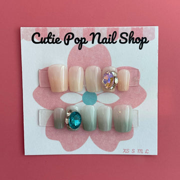 Pastel Gem press‑on nails by CutiePop Nail Shop featuring short, square-shaped nails in soft pastel hues—including blush pinks, mint green, and lavender—adorned with oversized iridescent gem embellishments and subtle metallic accents. Includes a reusable set with adhesive tabs, a mini nail file, and a cuticle stick, offering a sweet, dreamy manicure with a touch of luxe sparkle.