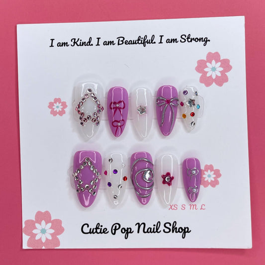 Purple Glamour press-on nails by CutiePop Nail Shop featuring medium-length almond-shaped nails in a mix of deep purple and crisp white tones. Accented with colorful gem clusters, embossed ribbon, star, and flower motifs, each nail is finished with a glossy shine for a chic and feminine glam look.