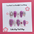 Purple Glamour press-on nails by CutiePop Nail Shop featuring medium-length almond-shaped nails in a mix of deep purple and crisp white tones. Accented with colorful gem clusters, embossed ribbon, star, and flower motifs, each nail is finished with a glossy shine for a chic and feminine glam look.

