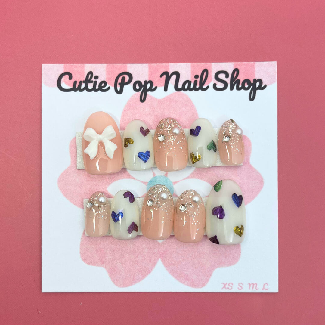 Rainbow Hearts press‑on nails by CutiePop Nail Shop featuring medium‑length round-shaped nails in a soft translucent pastel base, each adorned with hand-painted rainbow-colored heart motifs against a dreamy glossy finish. The nails come as a reusable kit—including ten handmade nails, jelly adhesive tabs, a mini nail file, and a cuticle stick—perfect for a playful yet elegant kaleidoscopic manicure.