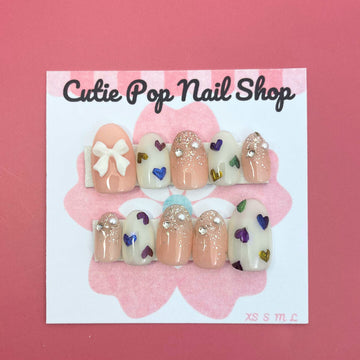 Rainbow Hearts press‑on nails by CutiePop Nail Shop featuring medium‑length round-shaped nails in a soft translucent pastel base, each adorned with hand-painted rainbow-colored heart motifs against a dreamy glossy finish. The nails come as a reusable kit—including ten handmade nails, jelly adhesive tabs, a mini nail file, and a cuticle stick—perfect for a playful yet elegant kaleidoscopic manicure.