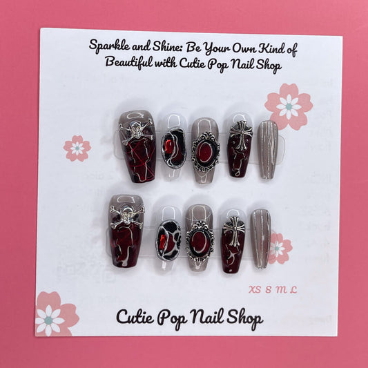Rebel Chic press‑on nails by CutiePop Nail Shop featuring a medium‑length coffin-shaped set combining glossy jet-black, deep blood-red, and transparent nails accented with edgy skull motifs, mirrored stone embellishments, and stylized cross details—hand-painted and hand-sculpted for a bold, gothic-rock aesthetic.
