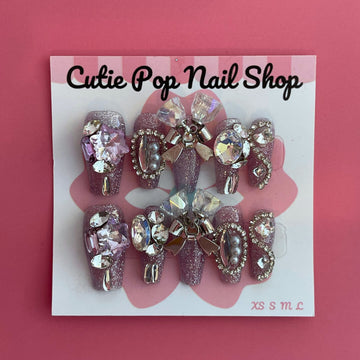 Saturn Bells press‑on nails by CutiePop Nail Shop featuring medium-length coffin‑shaped nails in a deep mauve or dusty rose tone, decorated with oversized iridescent gem clusters, metallic star and planet charms, and delicate glitter accents for a celestial, galaxy‑inspired aesthetic. Includes a set of ten reusable nails, adhesive tabs, mini nail file, and cuticle stick—perfect for dreamy cosmic glam.