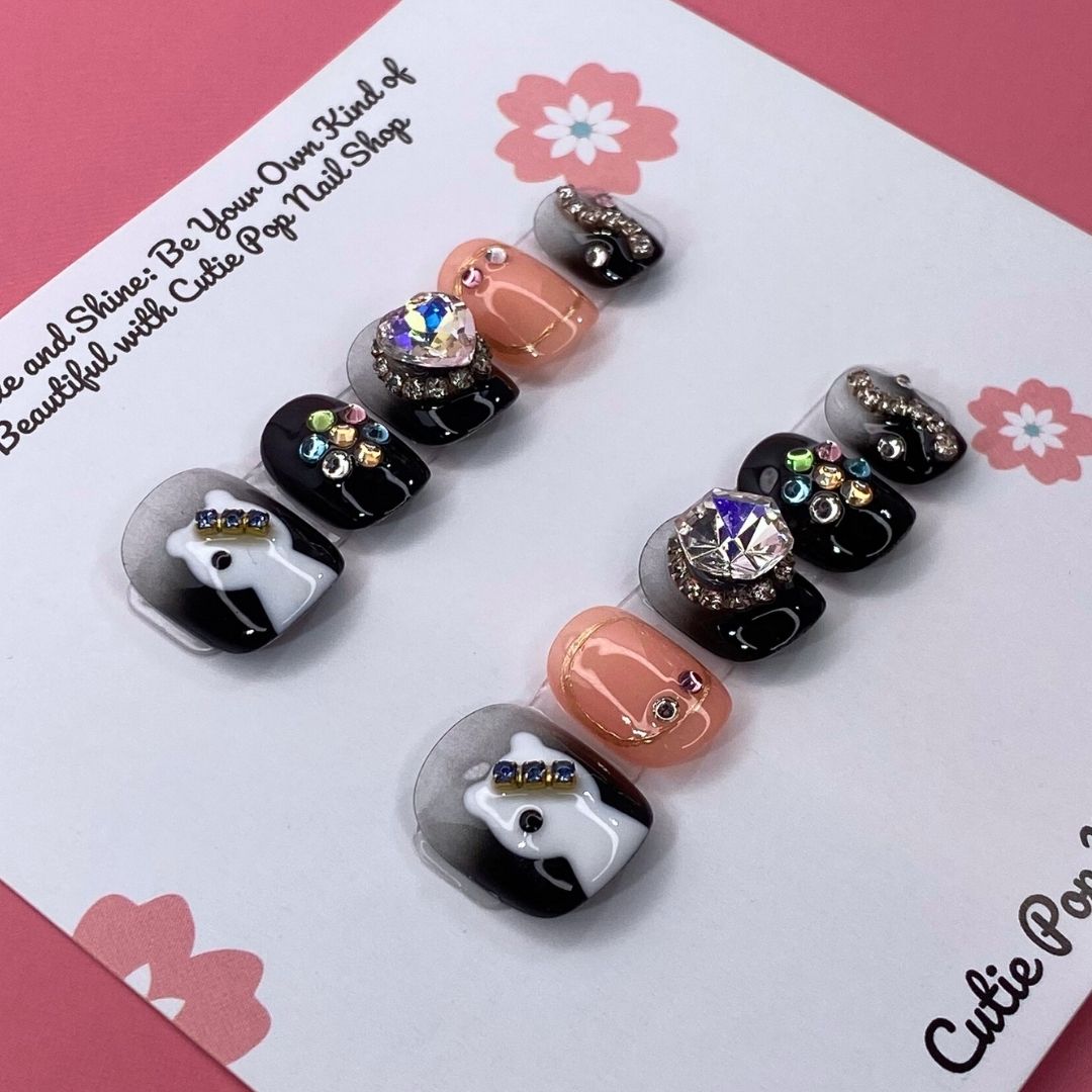Spooky Sparkle press-on nails by CutiePop Nail Shop featuring short square-shaped nails in a Halloween-inspired black and purple palette. Decorated with glitter, shimmer details, and cute spooky accents like black cats and ghostly elements, these nails add a fun, festive twist to your October look. Comes as a reusable kit with 10 handmade nails, jelly adhesive tabs, a mini nail file, and a cuticle stick for easy, mess-free application.