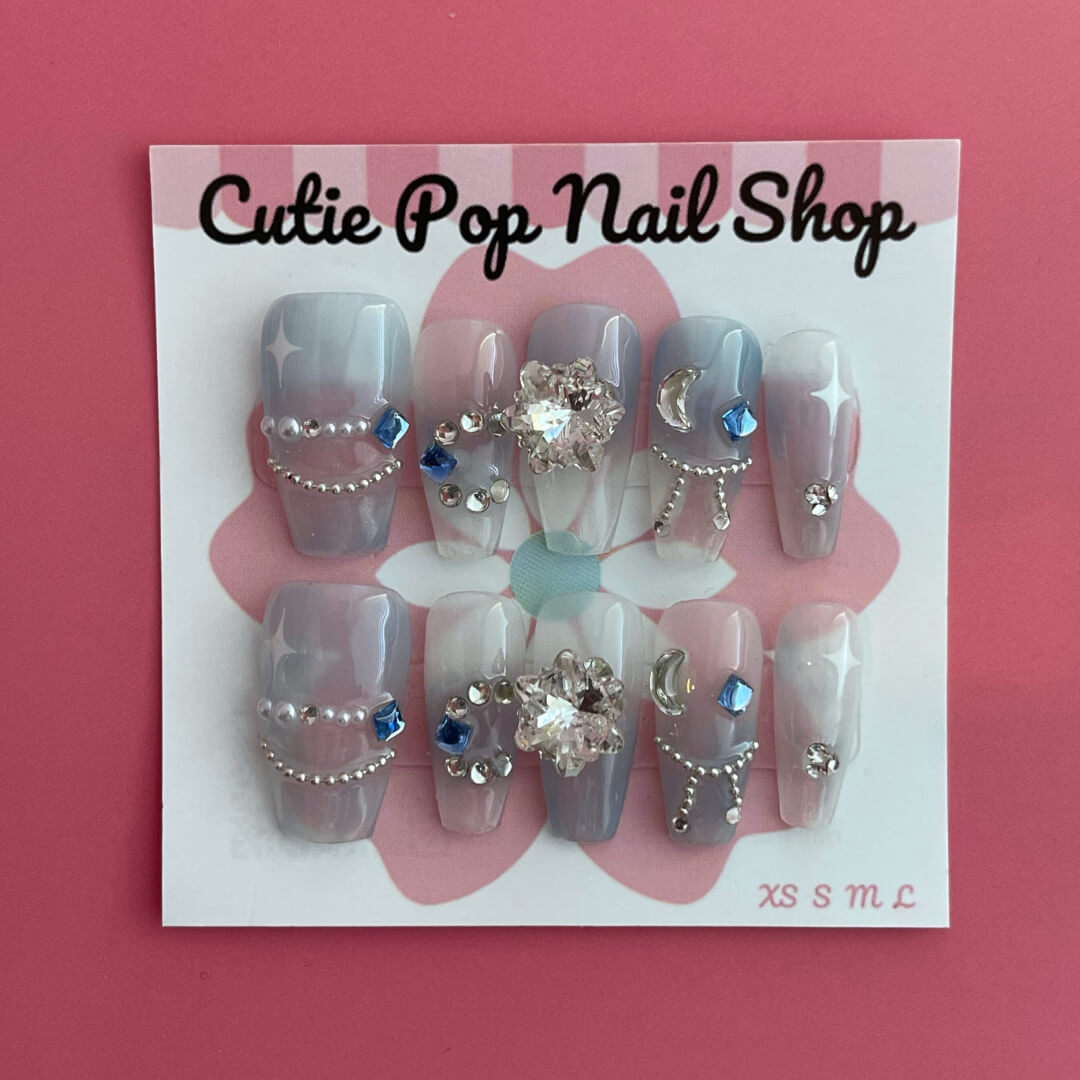 Star Gem (Blue) press on nails by CutiePop Nail Shop featuring medium coffin shaped nails in a translucent pale blue base adorned with sparkling silver star shaped gems and multicolored rhinestones for a celestial, glam star aesthetic. Includes a reusable adhesive tab set, a mini nail file, and a cuticle stick for full DIY application.