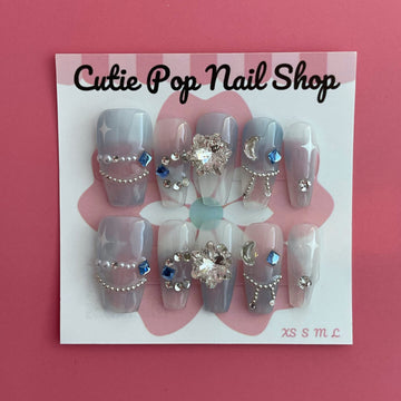 Star Gem (Blue) press on nails by CutiePop Nail Shop featuring medium coffin shaped nails in a translucent pale blue base adorned with sparkling silver star shaped gems and multicolored rhinestones for a celestial, glam star aesthetic. Includes a reusable adhesive tab set, a mini nail file, and a cuticle stick for full DIY application.