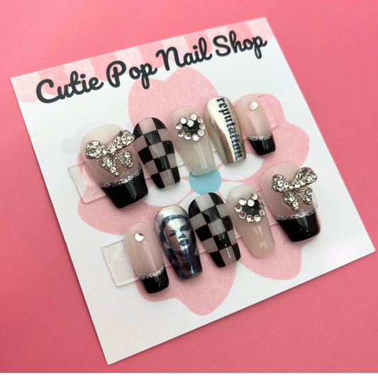 taylor swift inspired fake nails with checkered pattern and black french tip with bow on the thumb. The words "reputation" on a silver chrome nail, and taylor swift's face on another chrome nail. 